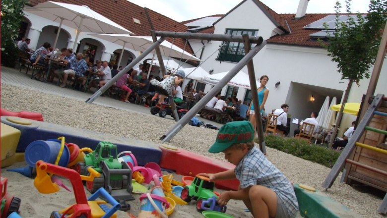 A child plays in a sandpit with colorful toys. In the background, people sit at tables under parasols.