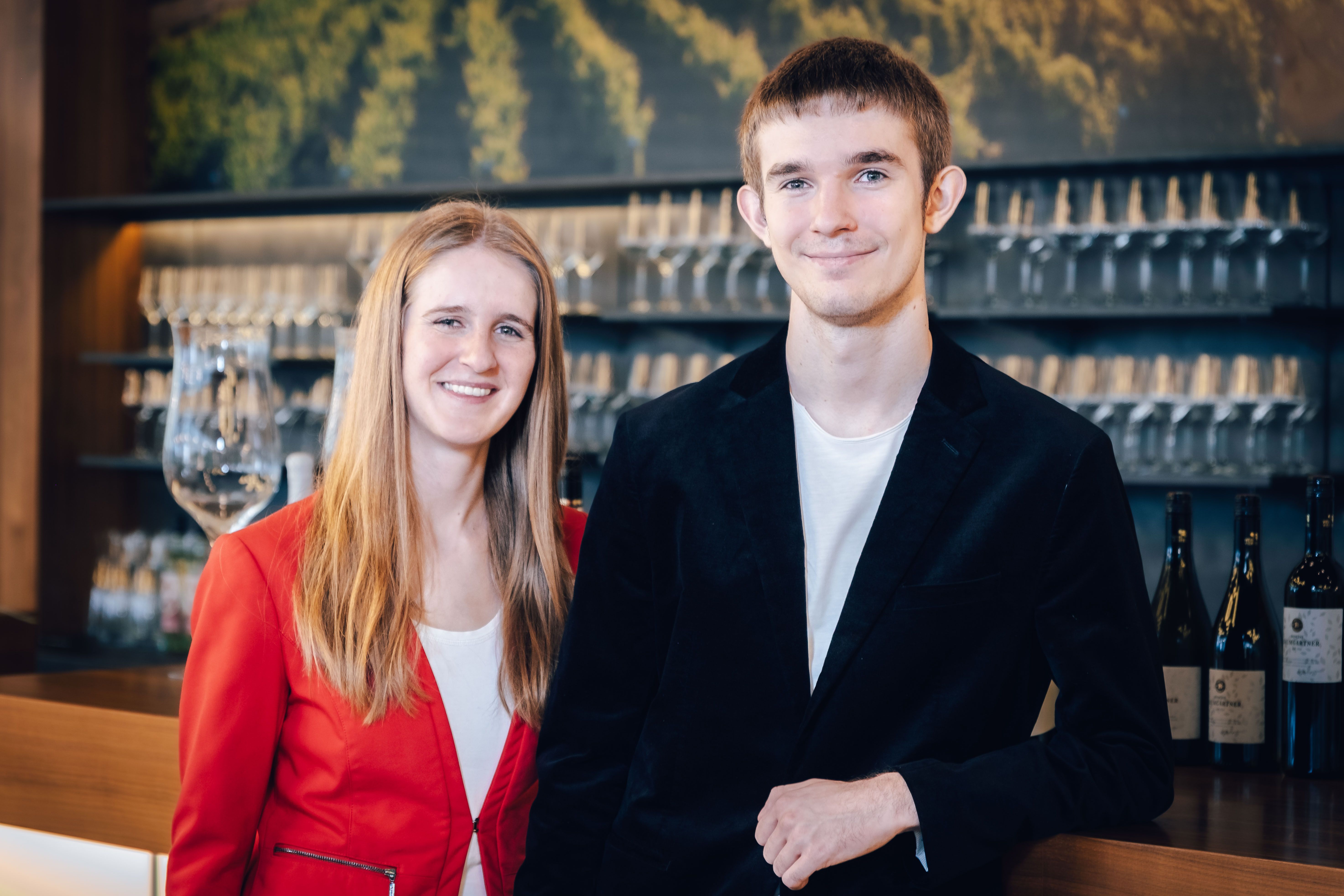 Katharina and Lorenz Baumgartner in elegant clothes in front of a shelf with wine glasses.