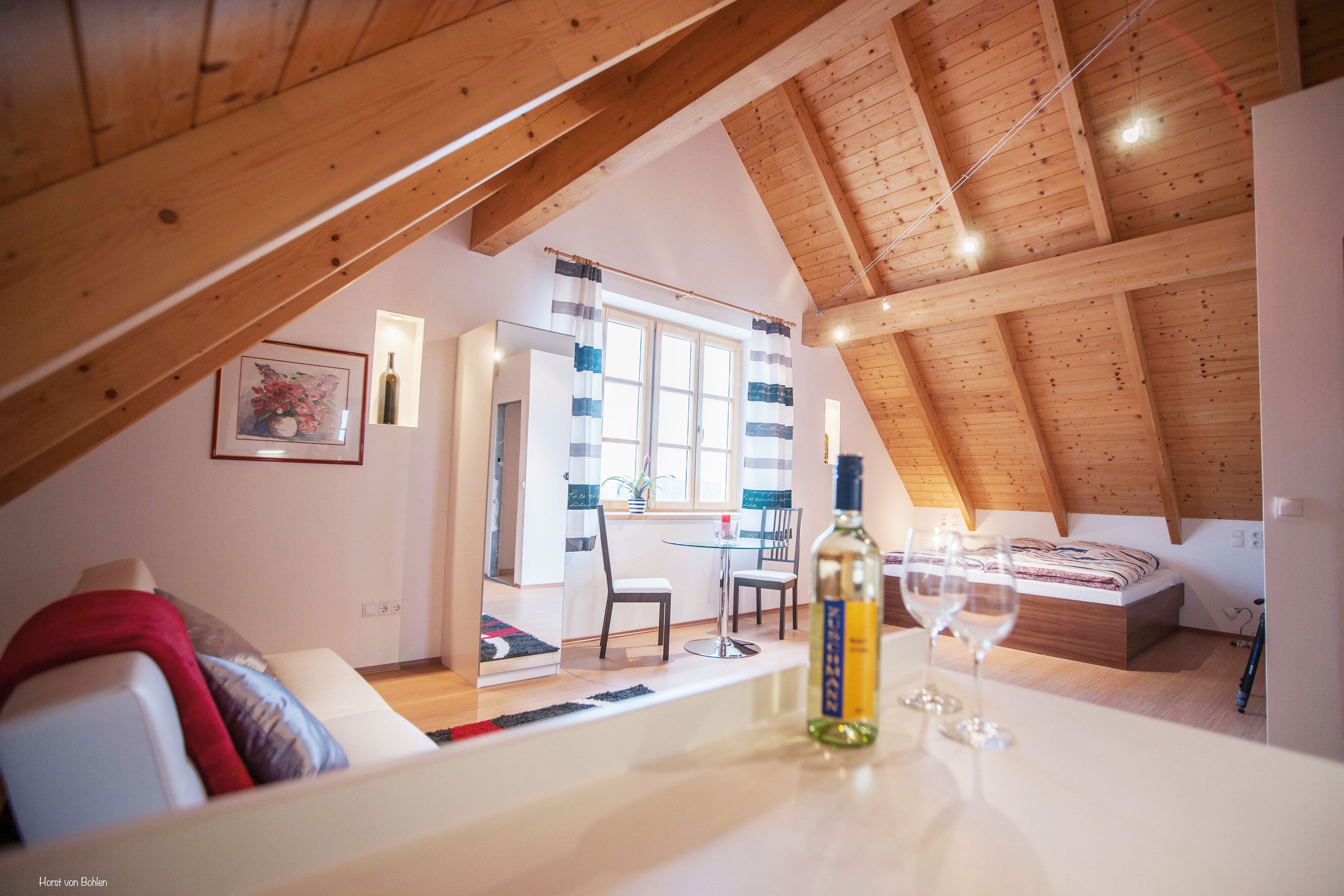 Cozy attic room with wooden beamed ceiling, bed, sofa, table and wine bottle.
