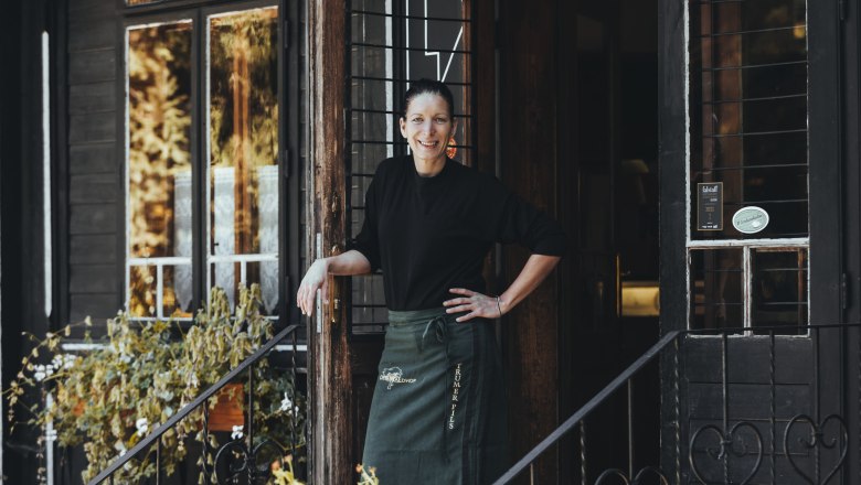 A woman stands smiling in the doorway of a rustic building with plants in the foreground.