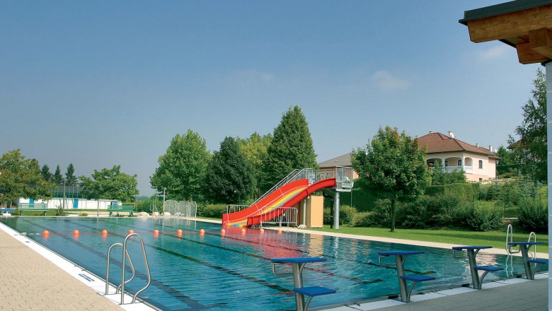 An outdoor pool with a swimming pool, a red slide and diving boards. Trees and a house can be seen in the background.