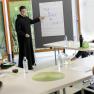 A seminar at Seitenstetten Abbey with a lecturer dressed as a monk pointing at a flipchart. Participants sit at tables.