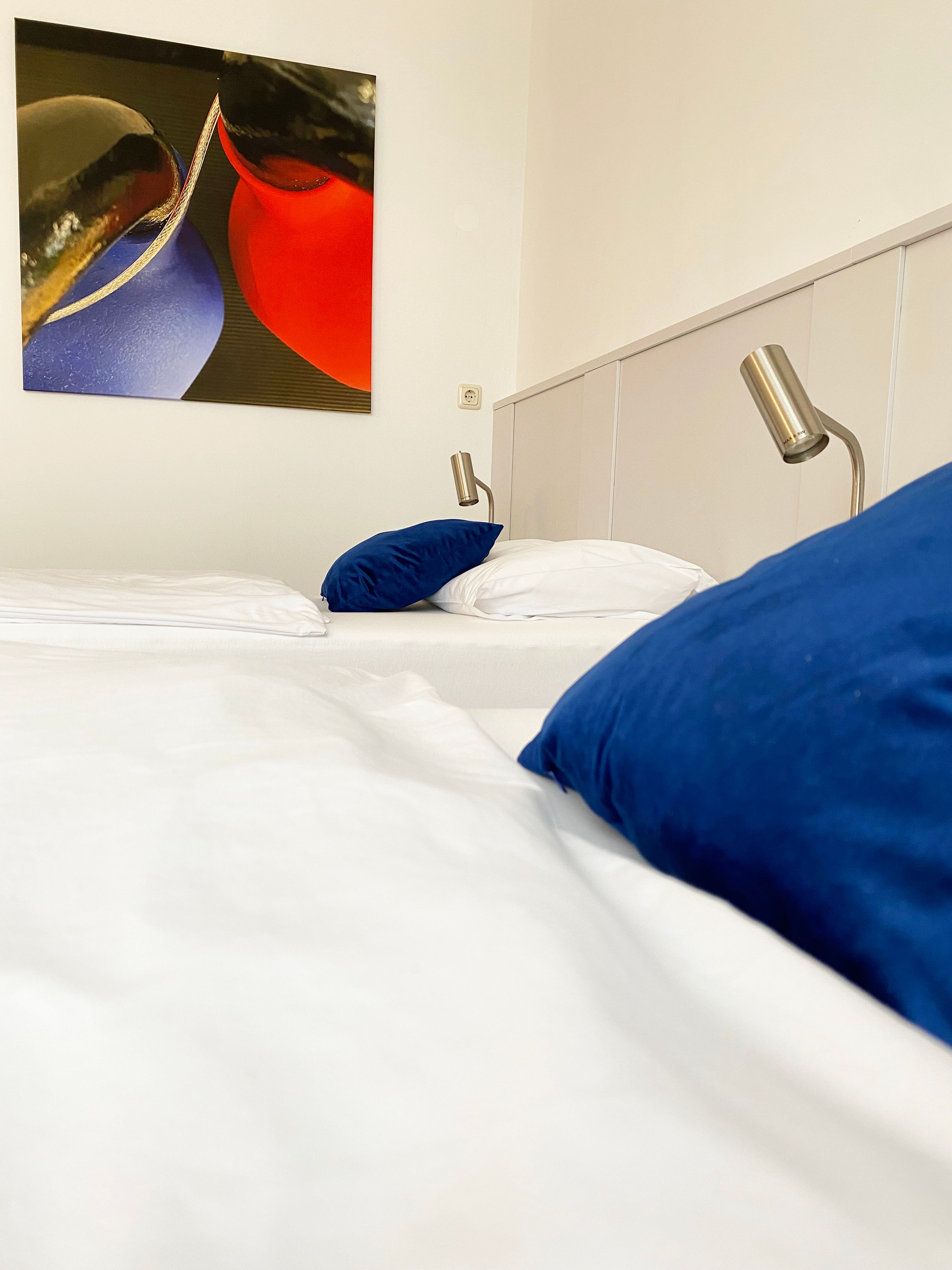 A guest room with a bed, blue cushions, reading lamps and an abstract painting on the wall.