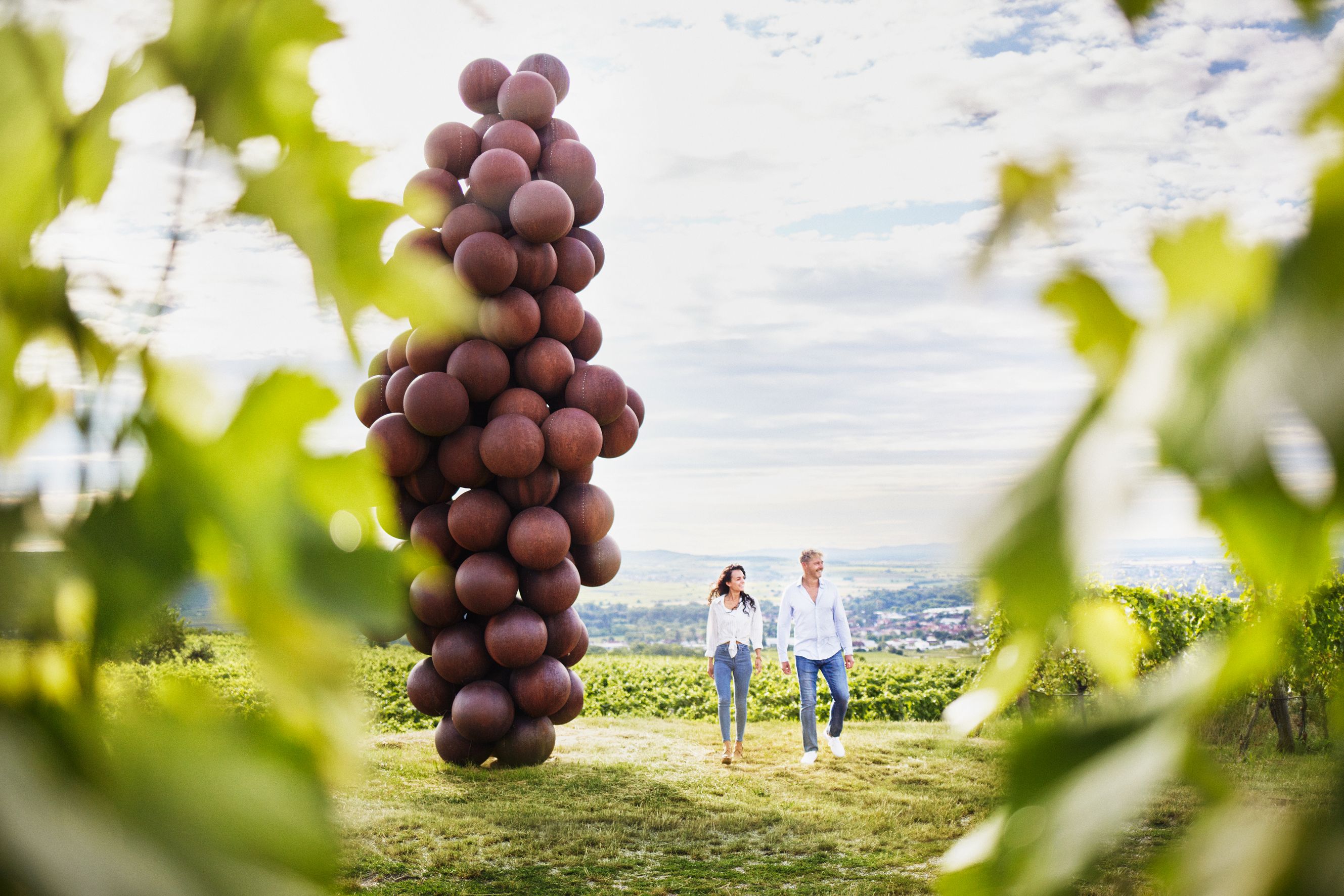 Two people walk past a large sculpture made of spherical elements, surrounded by vineyards.