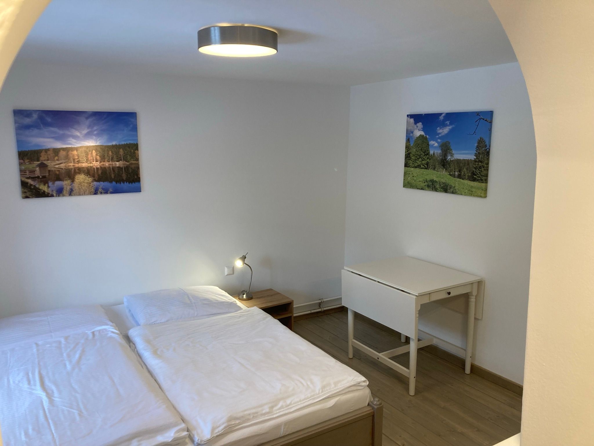 A simply furnished room with a double bed, a small table and two landscape paintings on the wall.