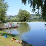 A quiet lake with a jetty and anglers, surrounded by green trees and hills in the background.