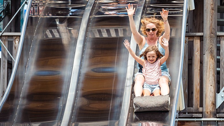 A woman and a child with raised arms sit sliding on a mat on a silver slide.