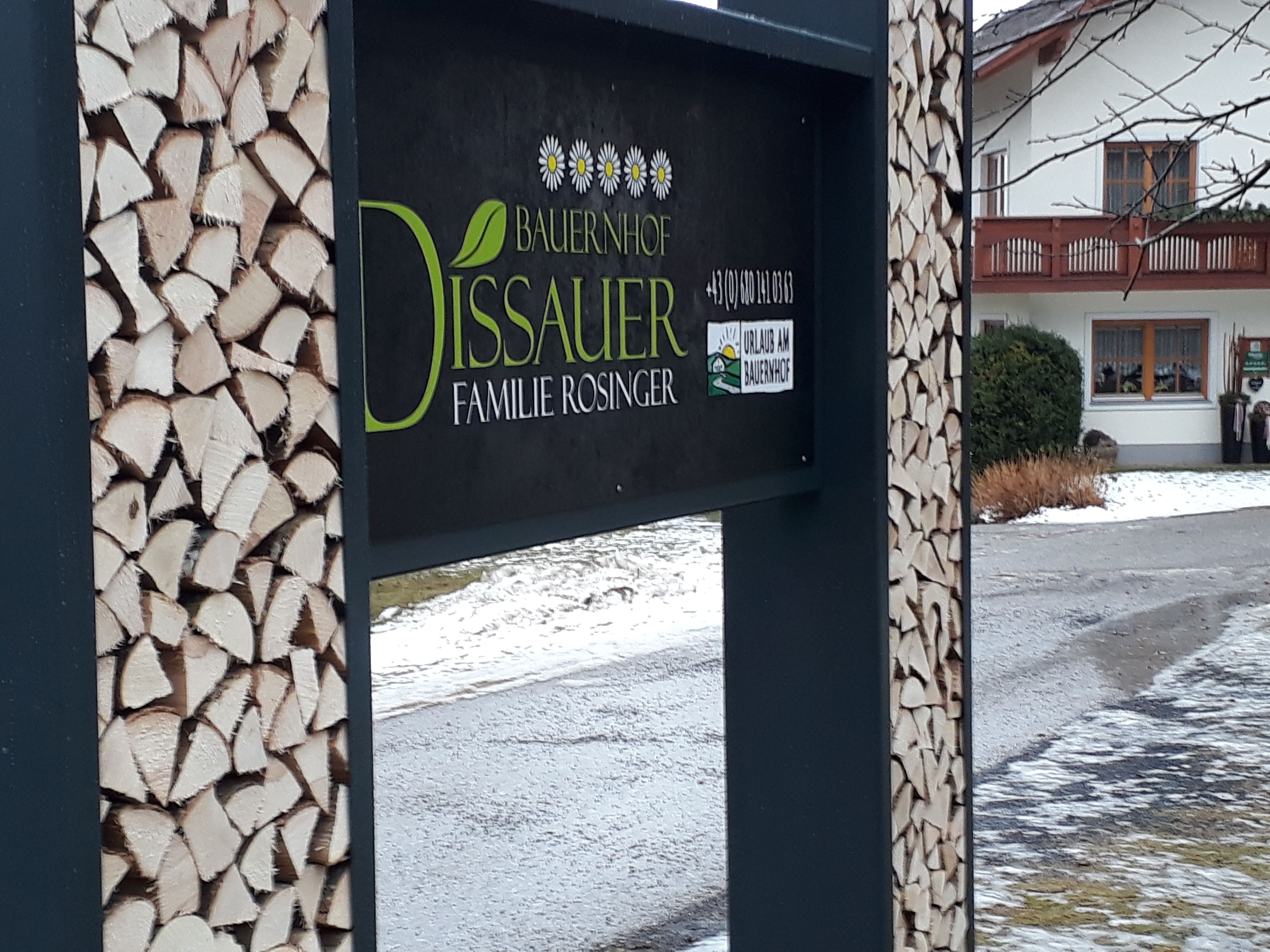Sign of a farm with piles of wood and a house in the background.
