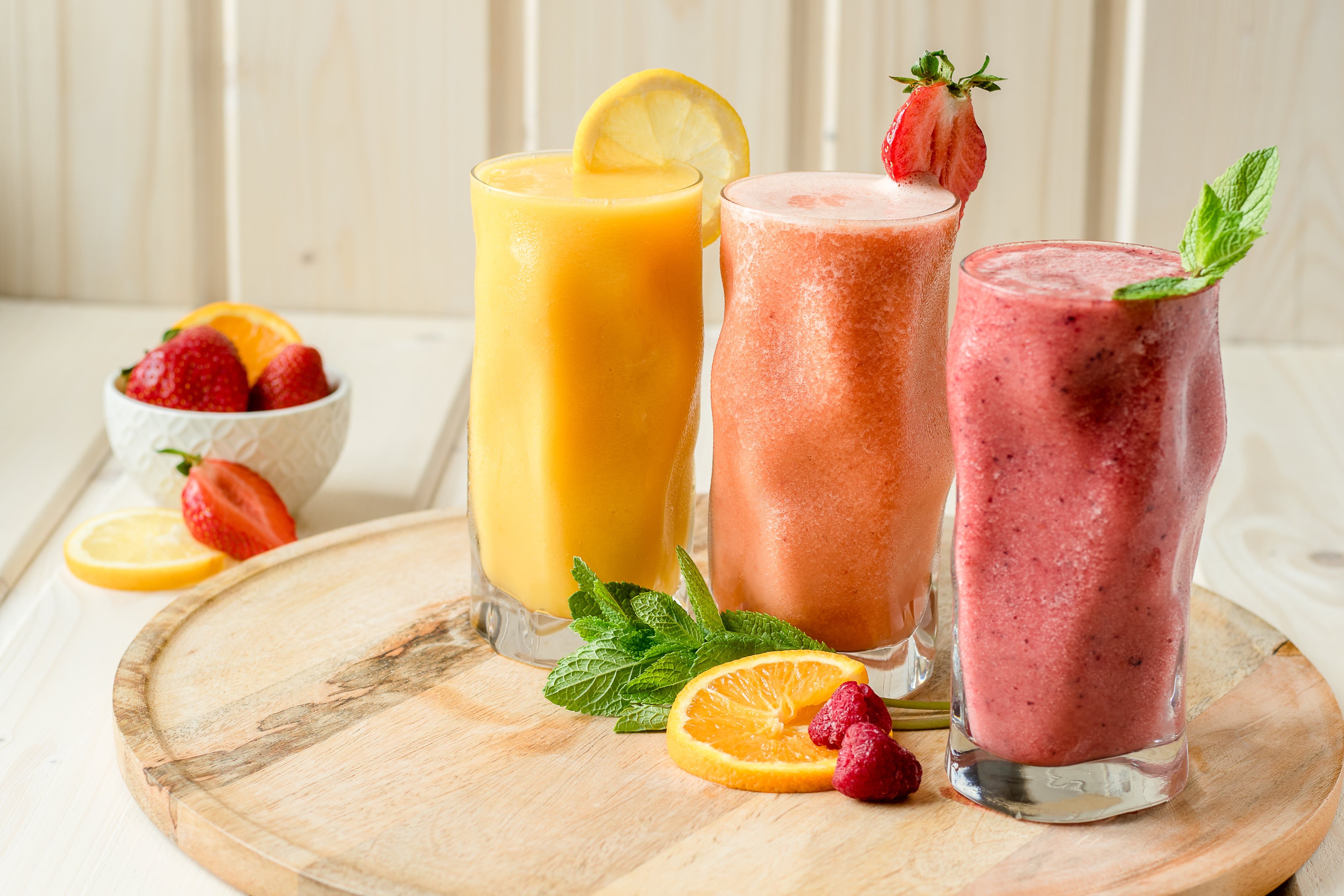 Three colorful smoothies in glasses decorated with fruit and mint on a wooden tray.