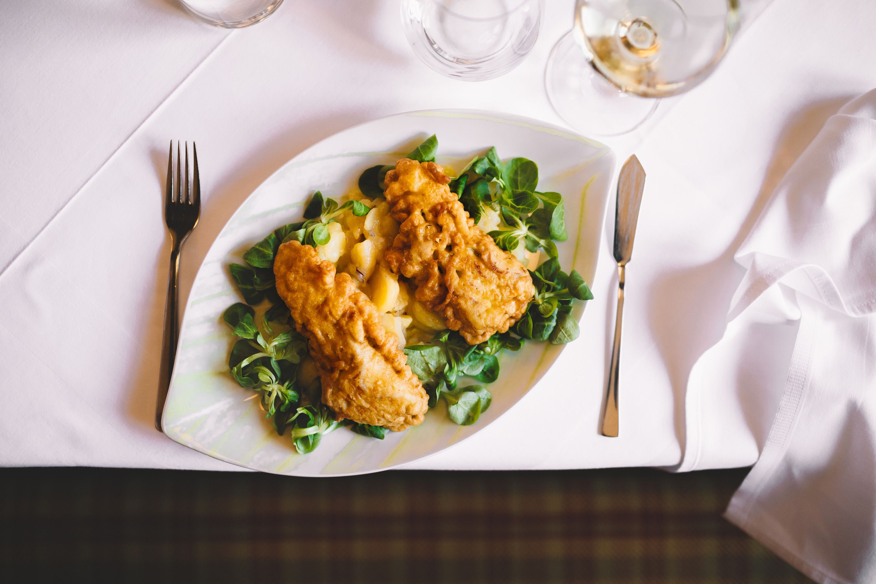 A plate of baked carp fillet on lamb's lettuce and potatoes, on a table set in white.