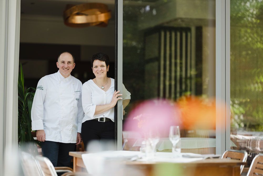 A man in chef's clothes and a woman stand smiling in an open doorway of a restaurant.