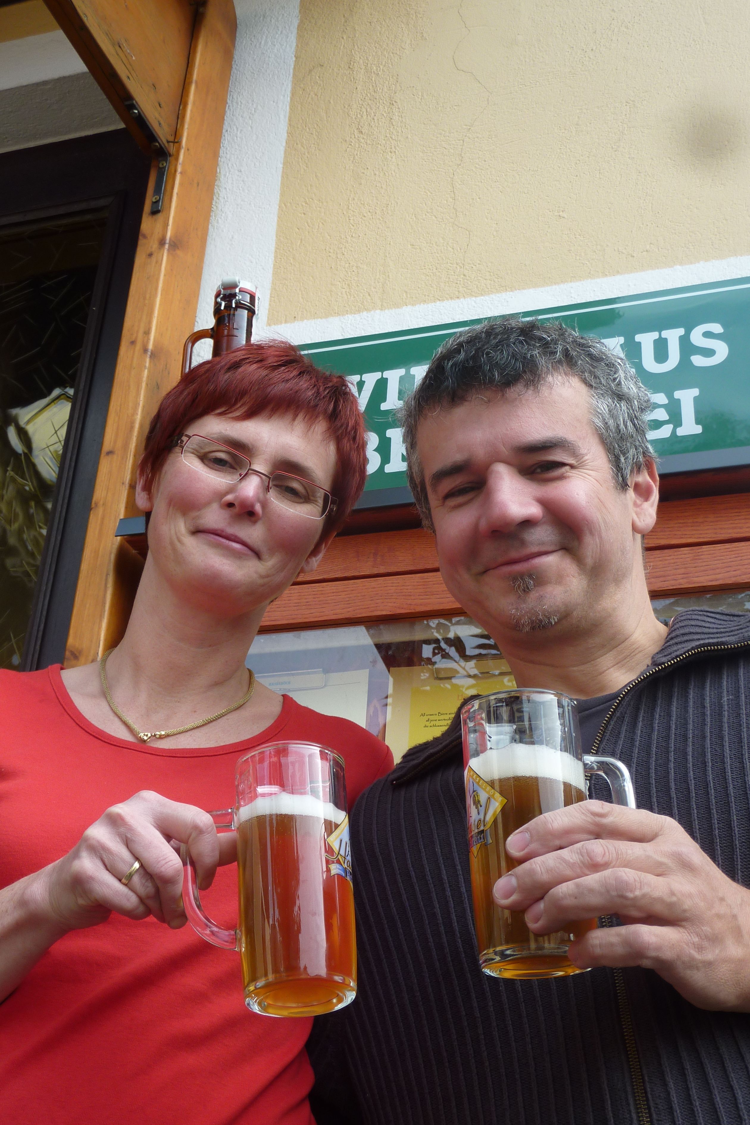 Two people clink glasses of beer in front of a sign for the Haselböck brewery.