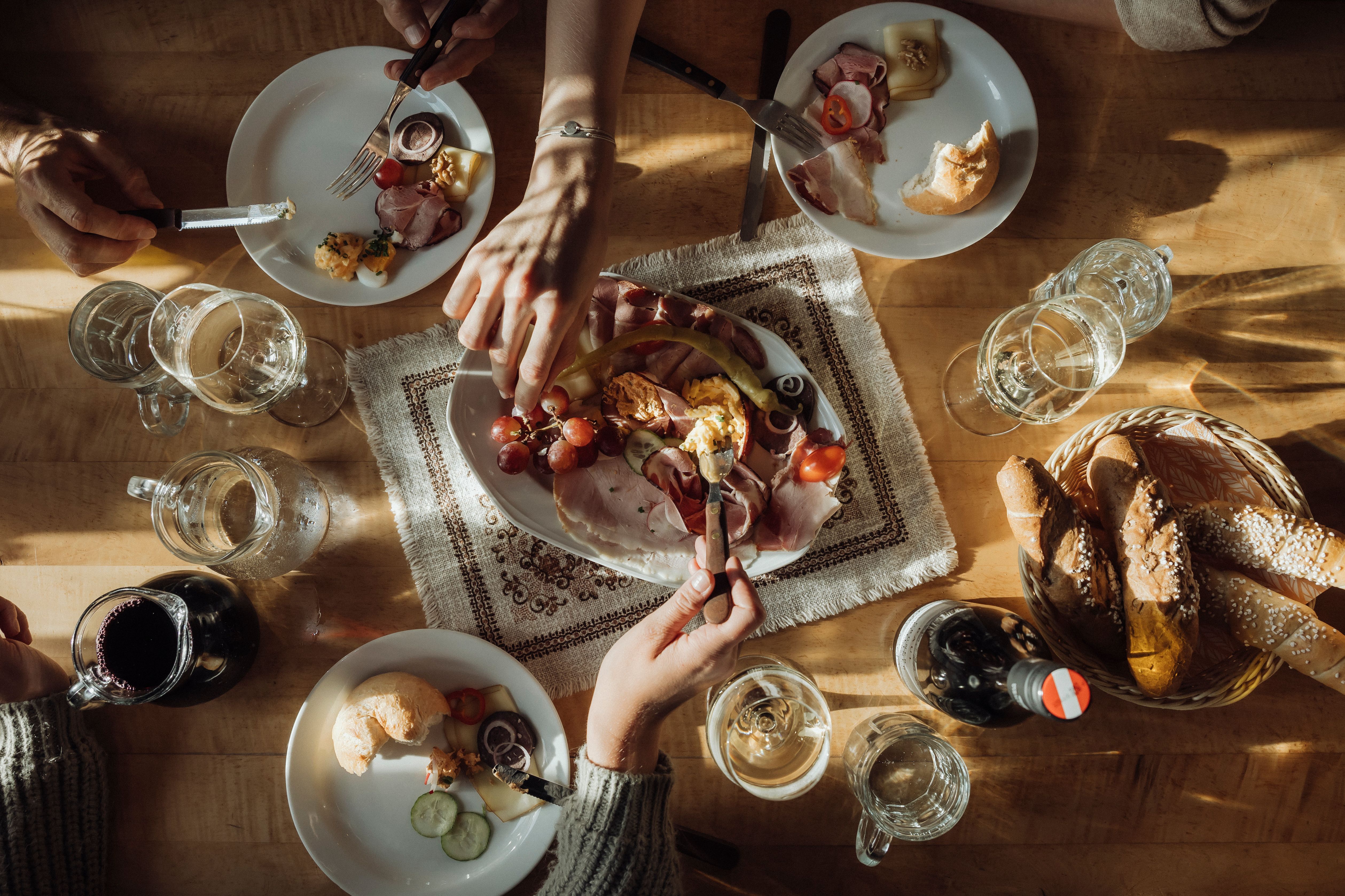 A table with a Heurigen snack consisting of bread, cold cuts, cheese and wine, surrounded by several hands.