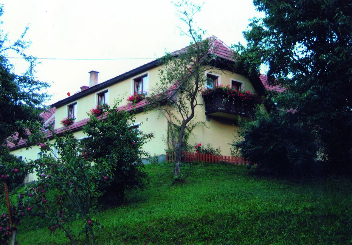 A yellow house with red roof tiles and flower boxes on the windows, surrounded by trees and a green slope.