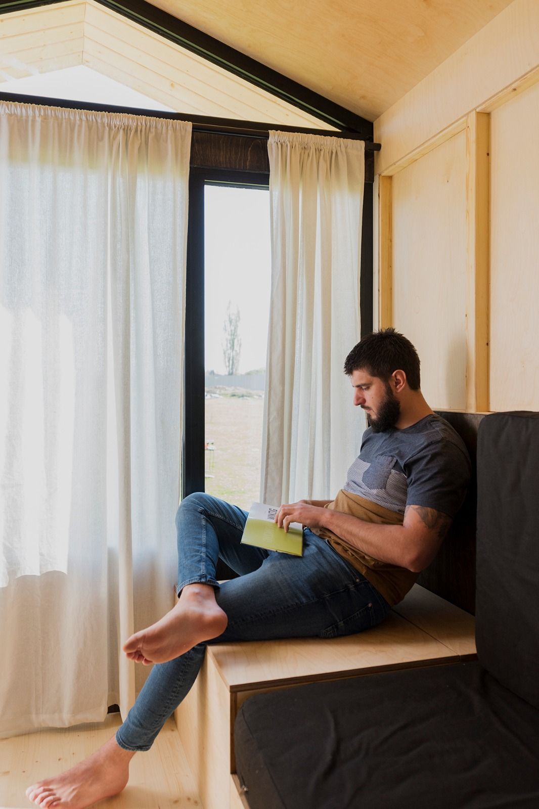 A man is sitting in a cozy corner of a wooden hut reading a book. Daylight is streaming in through the window.
