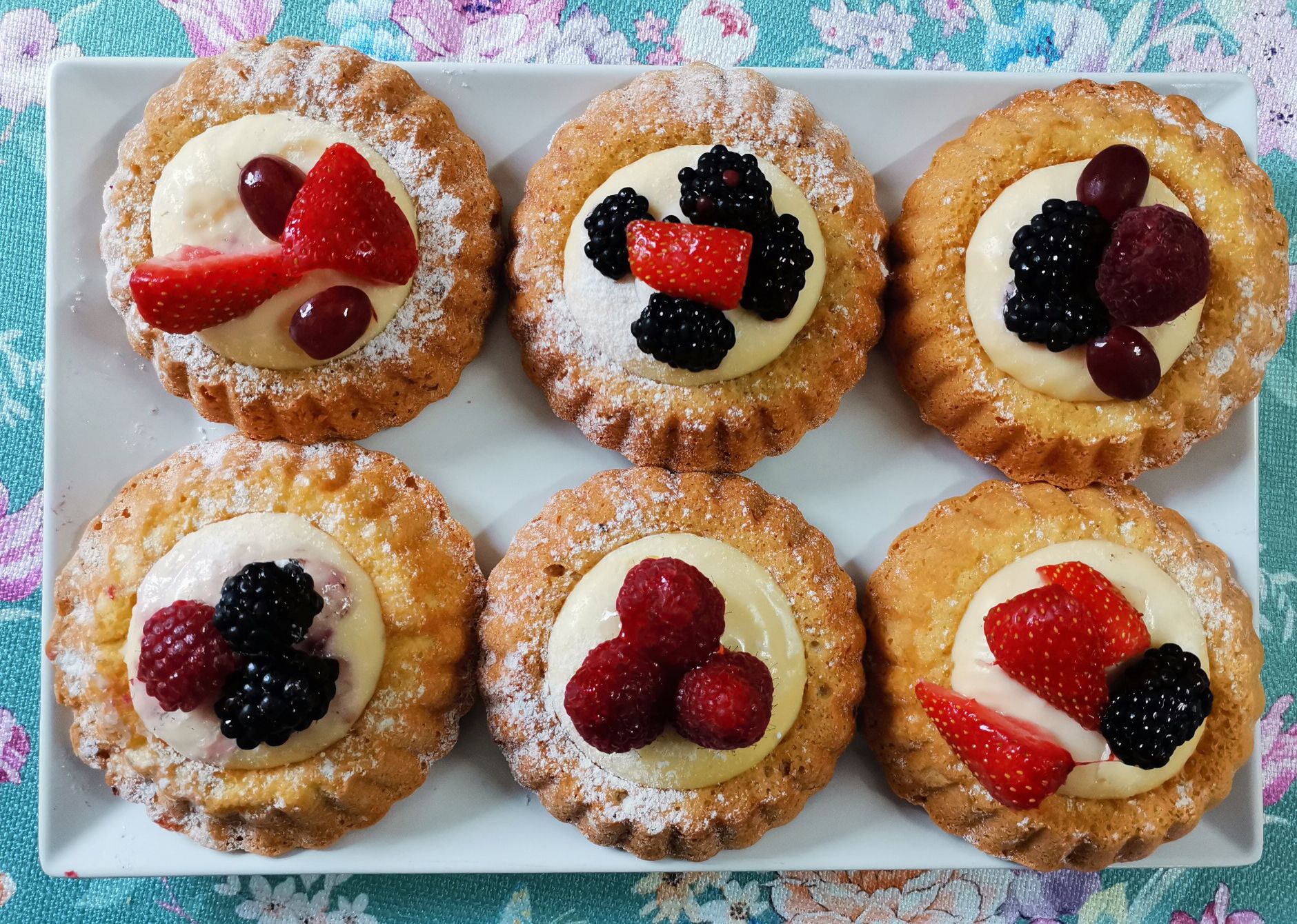 Six small fruit tarts on a white plate, decorated with berries and cream.
