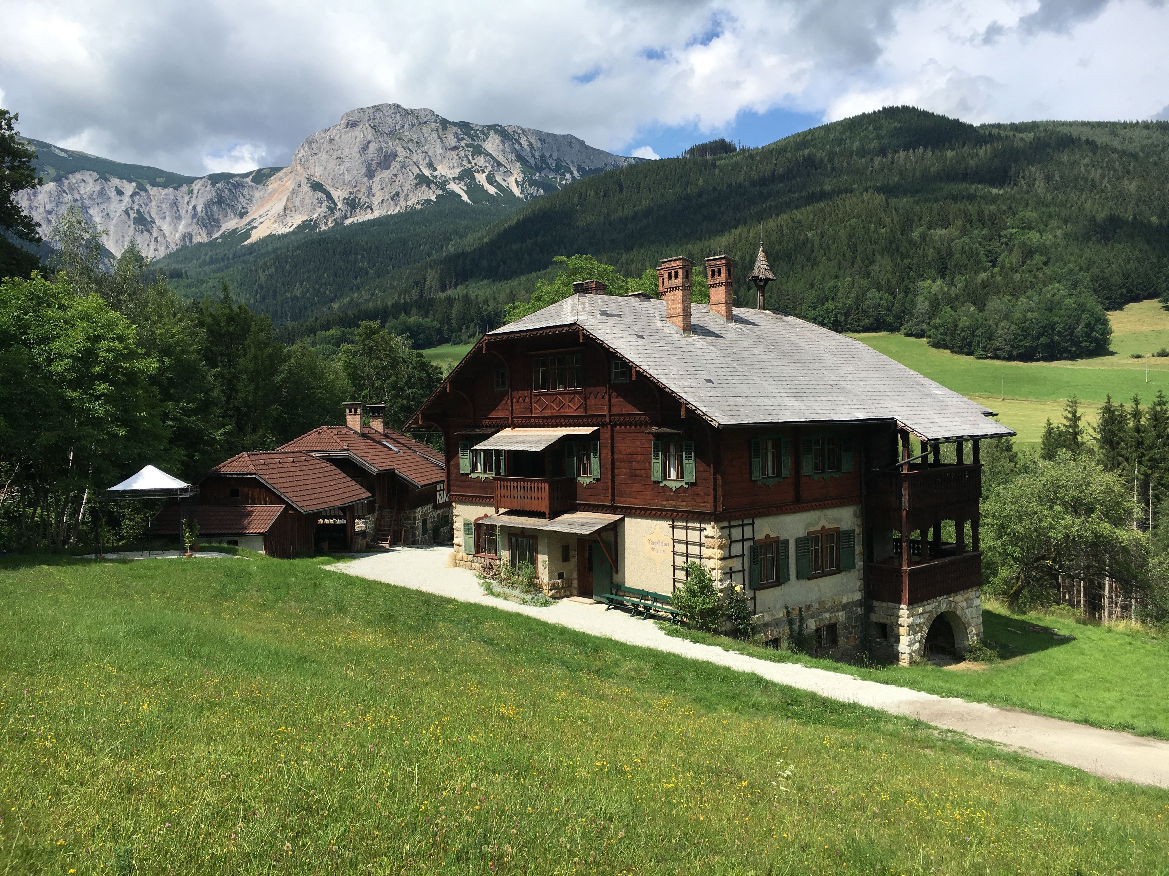 A traditional wooden house in a mountainous landscape with green meadows and wooded hills in the background.