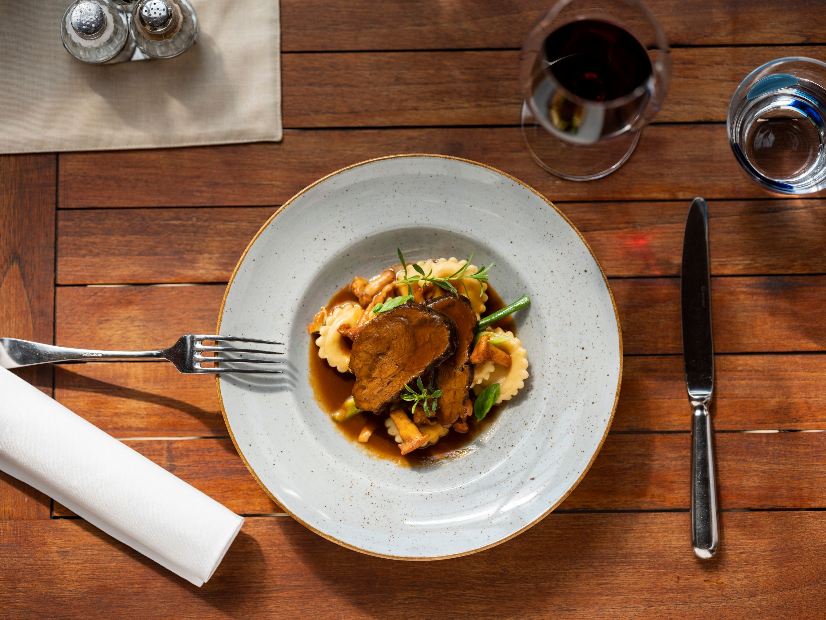 A plate of braised beef cheeks, ravioli and vegetables on a wooden table, with cutlery and a glass of red wine next to it.