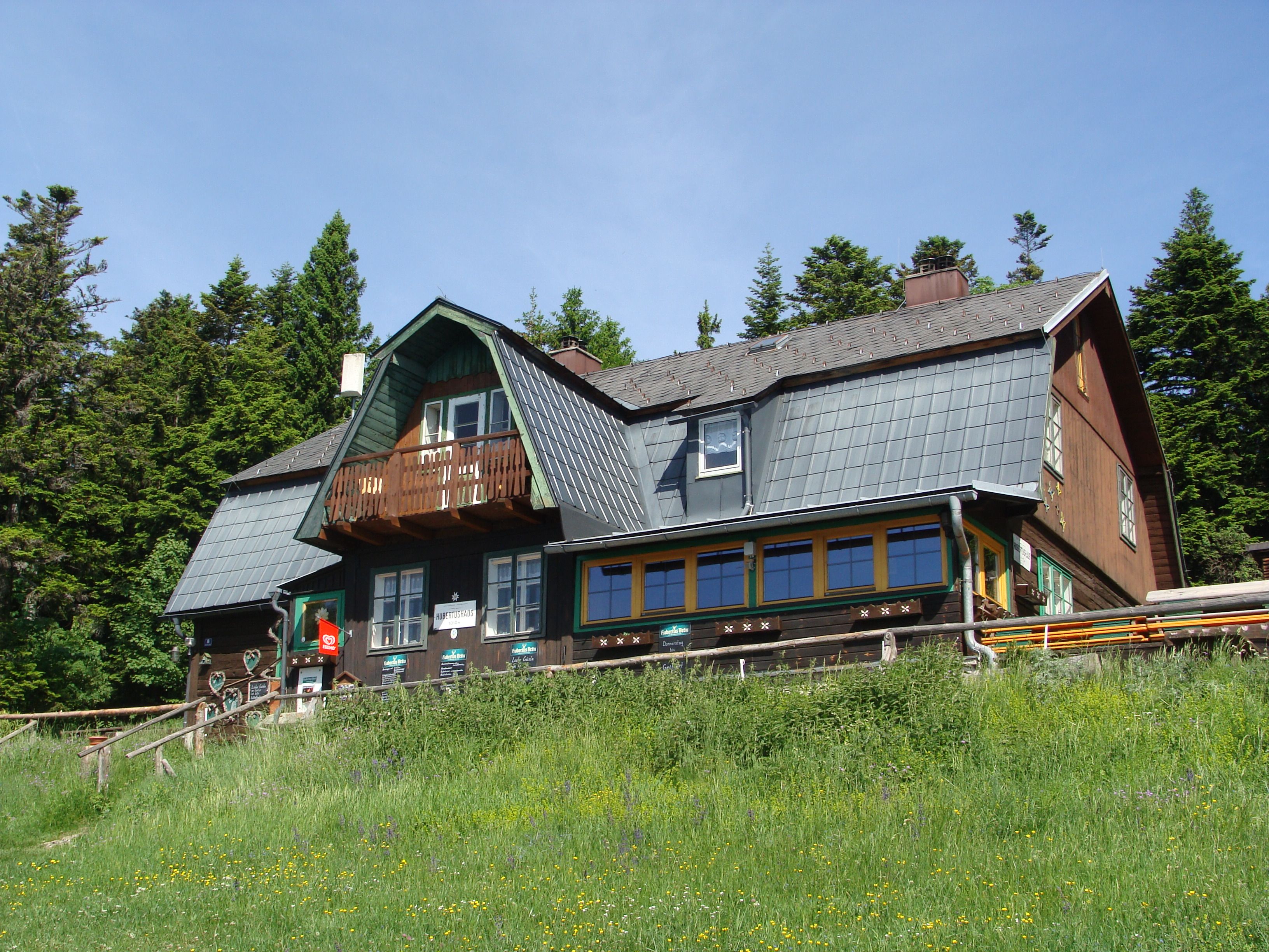 A traditional mountain house with a wooden façade and shingle roof, surrounded by trees and meadows.