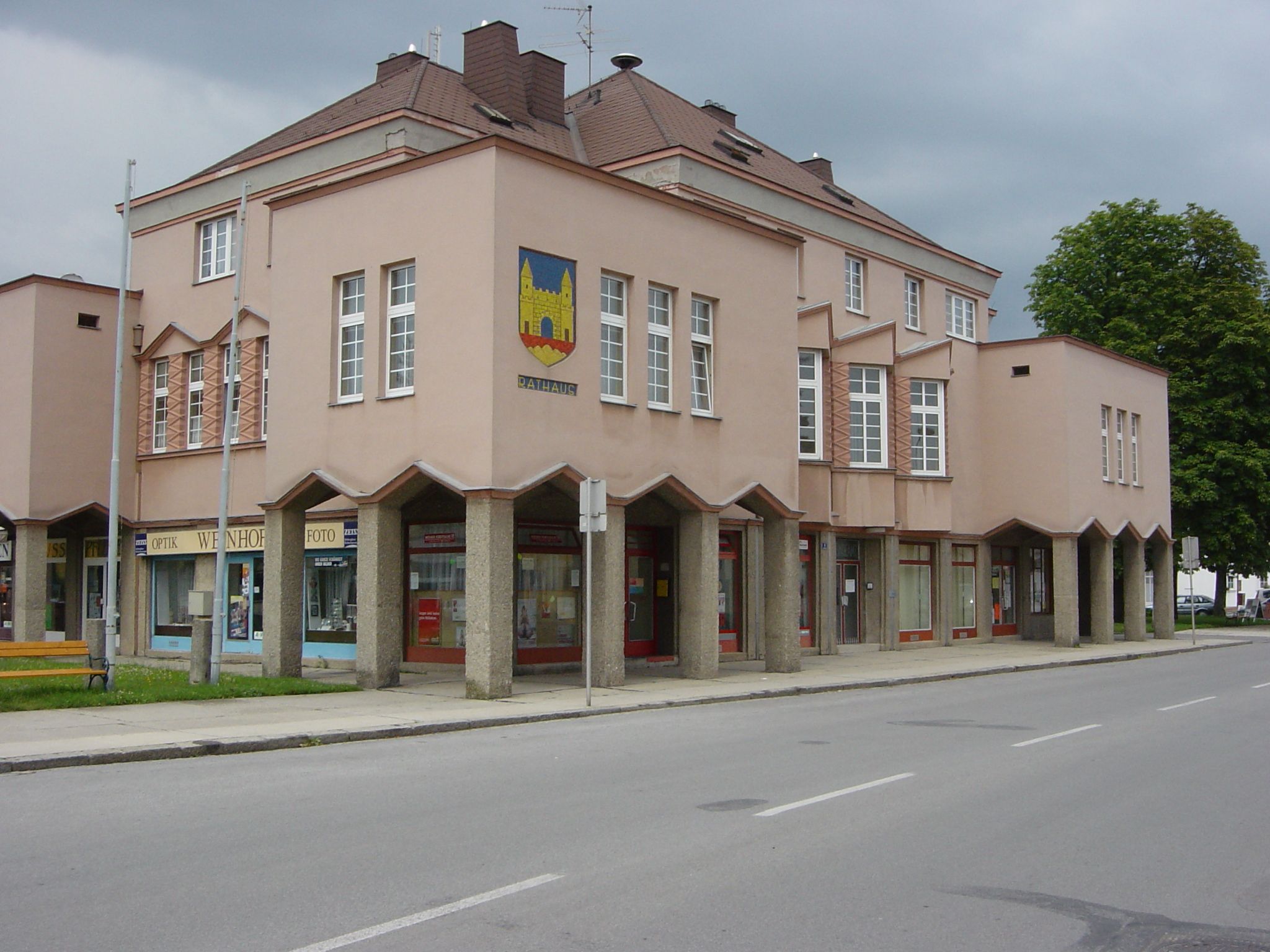 A modern town hall building with a pink façade and large windows. It has a coat of arms on the corner and stores on the first floor.