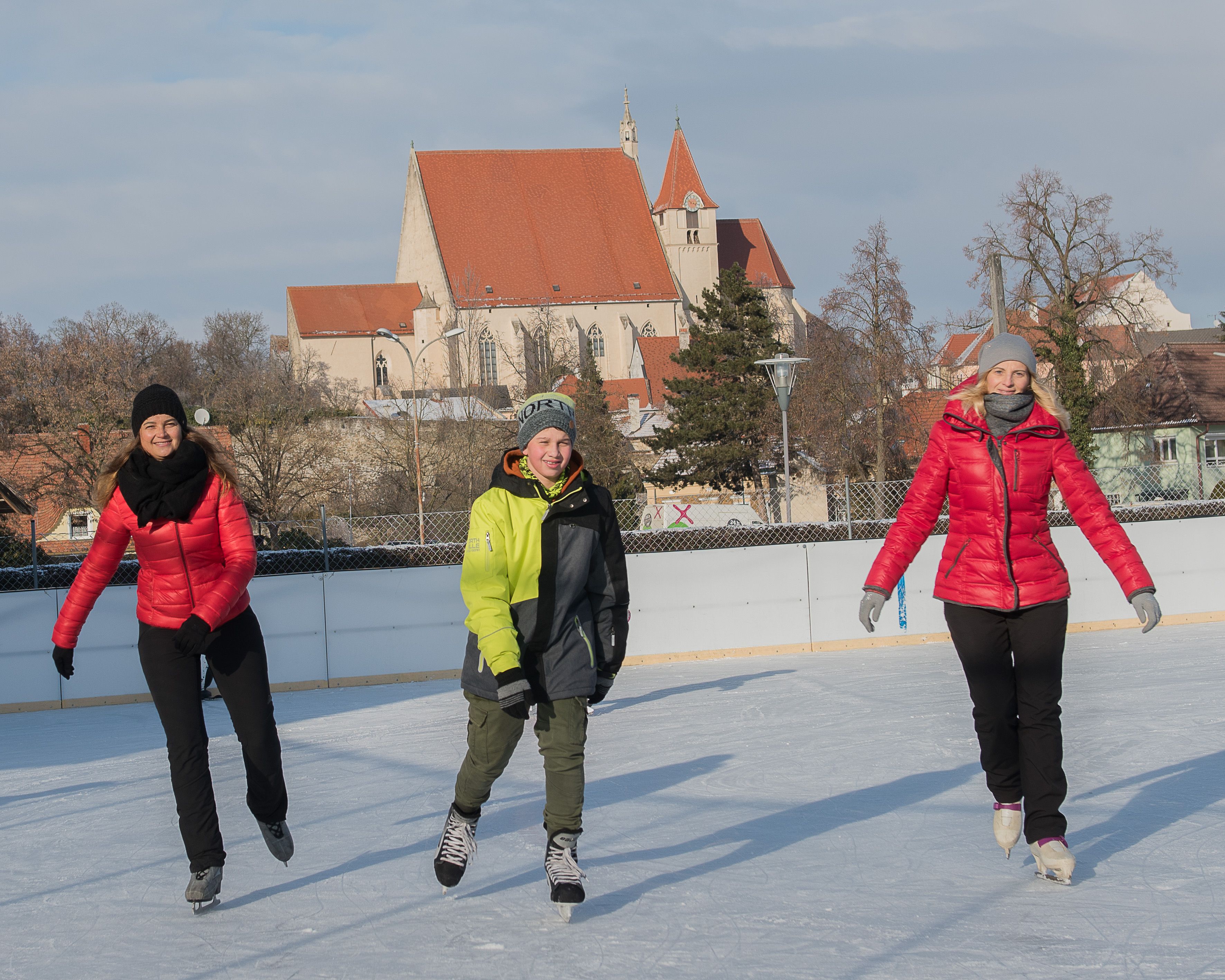 Three people skating on an ice rink with a church in the background.