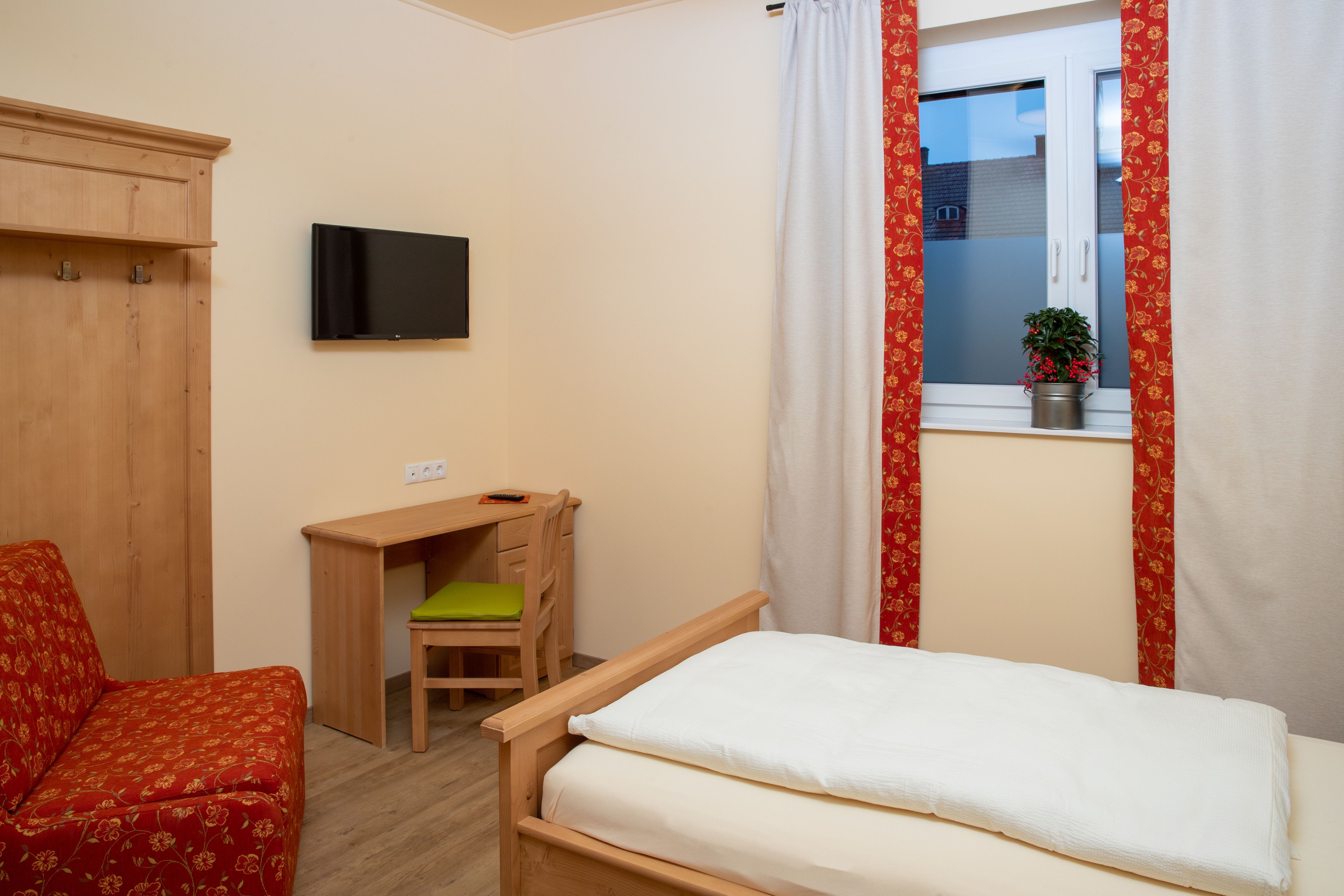 A cozy single room with wooden furniture, a bed, a desk and a TV on the wall. A window with curtains and a plant.