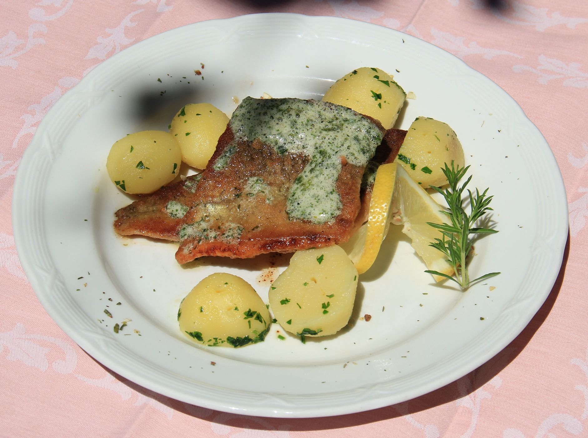 A plate of fried trout fillet, potatoes, lemon slices and herbs on a pink tablecloth.