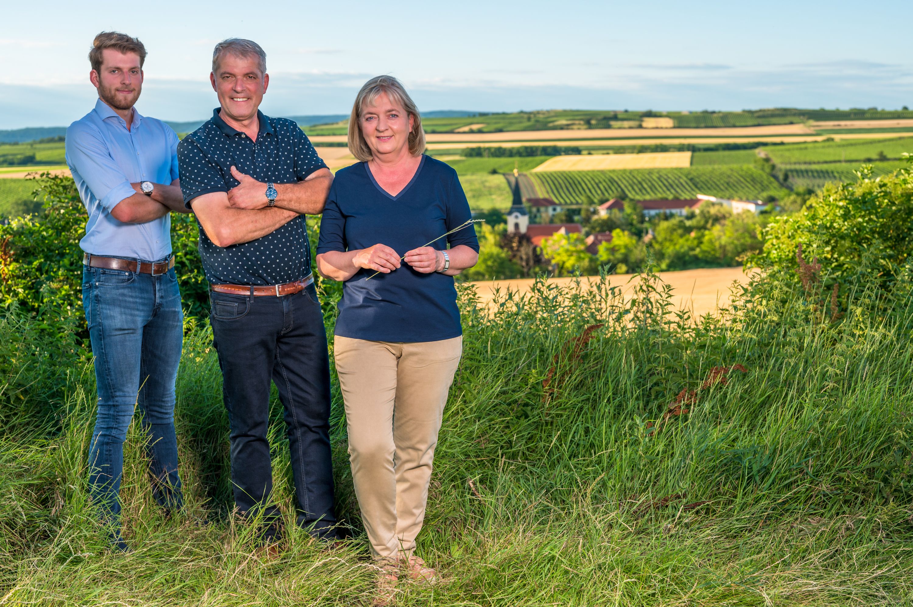 Three people are standing in a vineyard with a view of a rural landscape in the background.