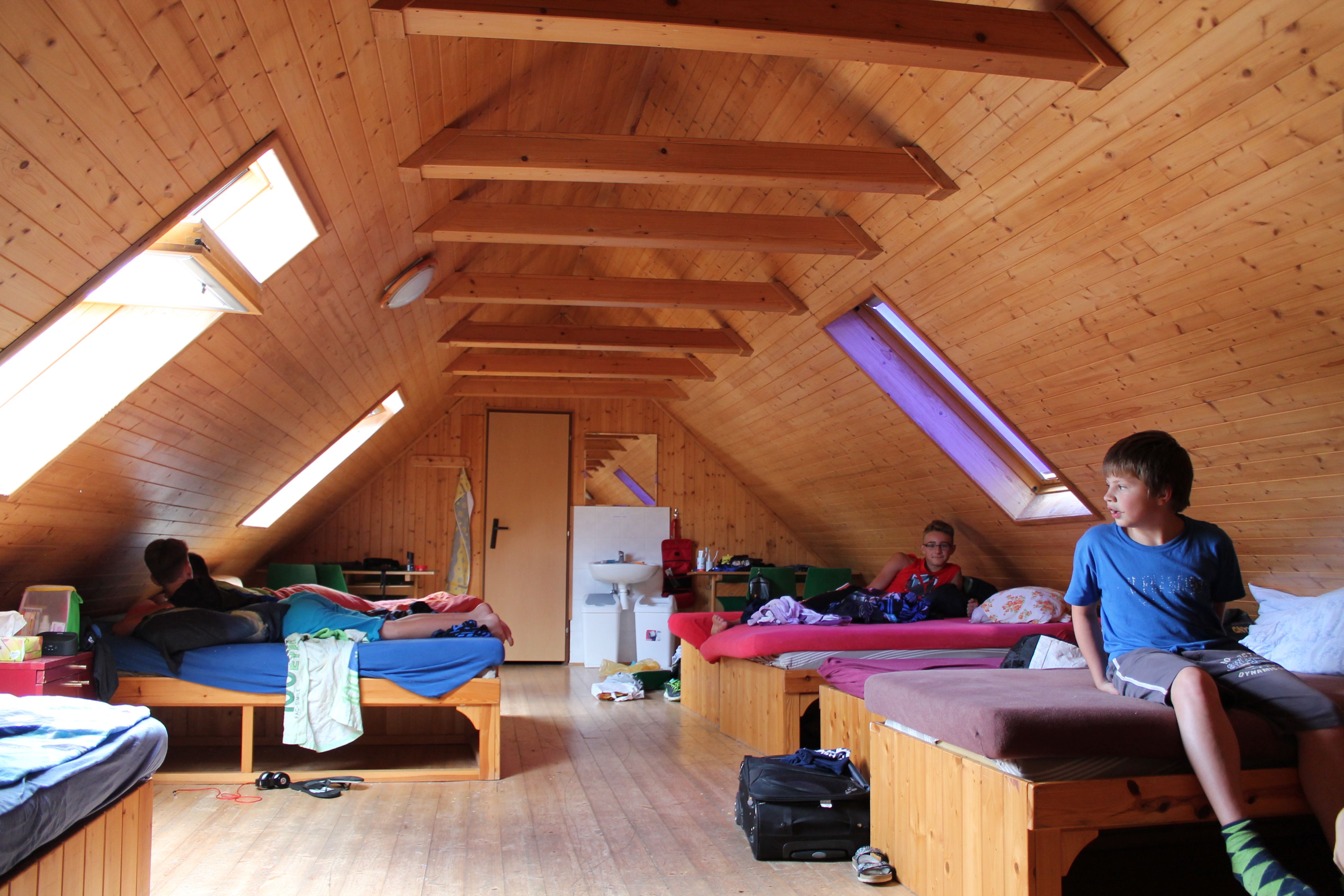 Attic room with wooden walls, several beds and skylights. Three people are in the room, one is sitting on a bed.