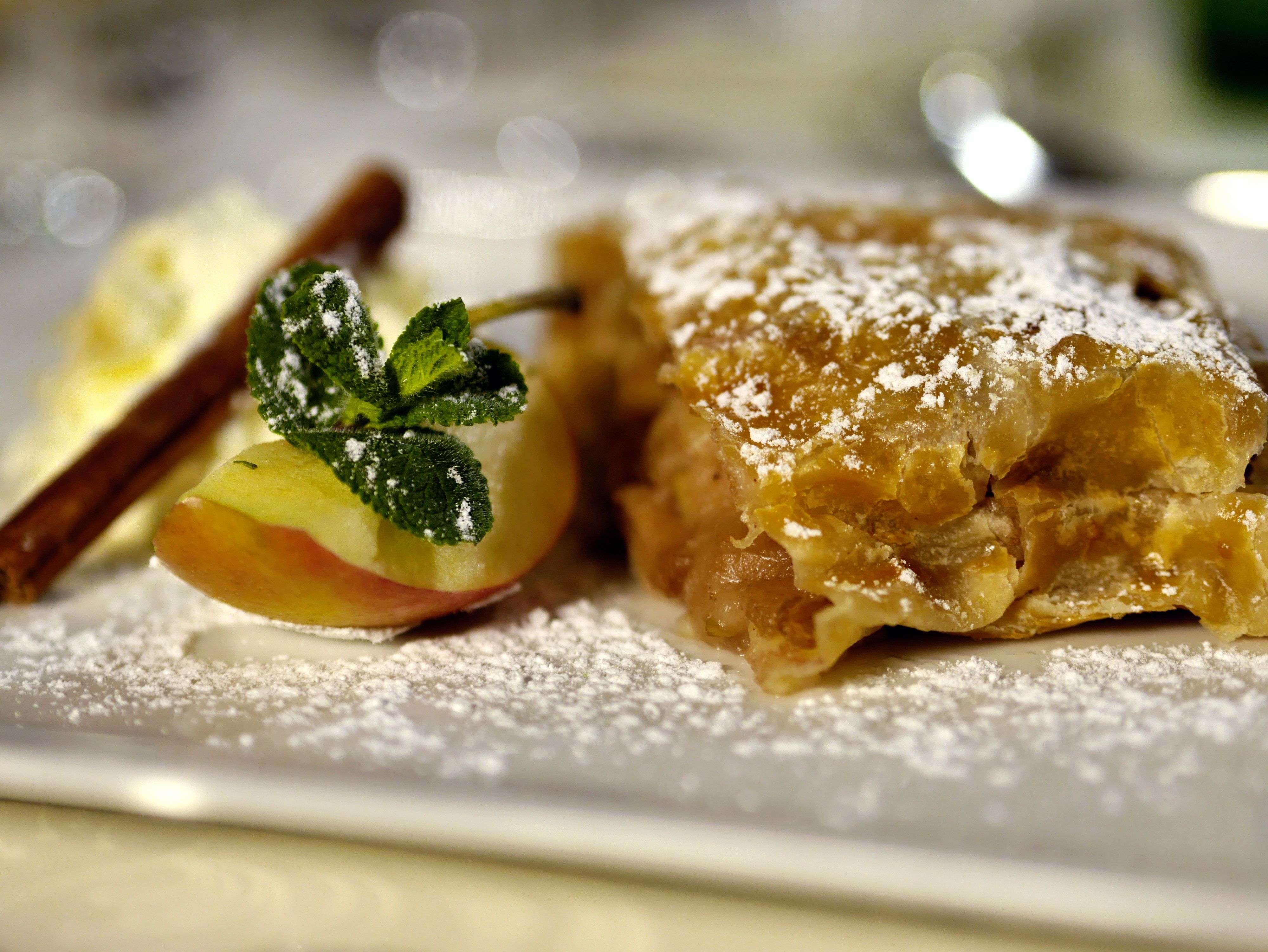 A piece of apple strudel with powdered sugar, apple slice, cinnamon stick and mint leaf on a plate.