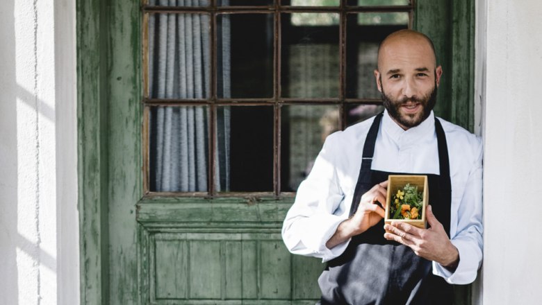A cook in a white uniform holds a small wooden box of herbs in front of a green door.