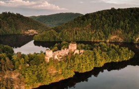 Aerial view of a ruined castle on a wooded peninsula surrounded by a calm lake.