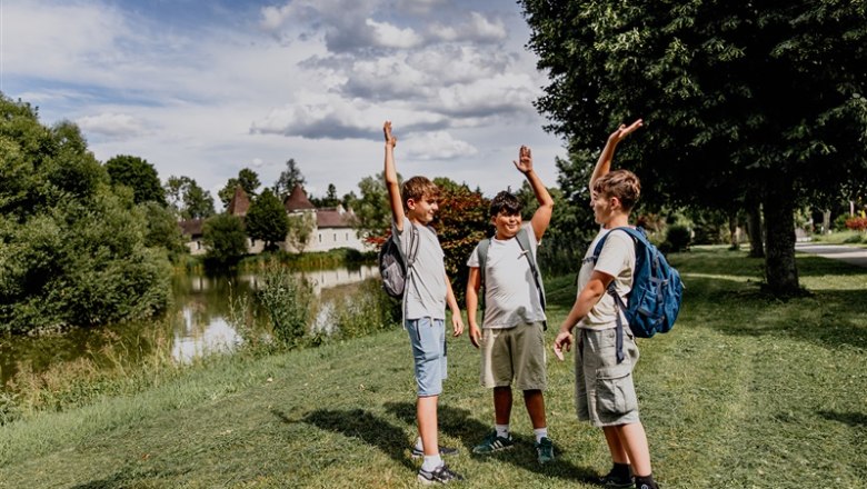 Three boys with rucksacks stand on a meadow next to a pond and raise their arms in the air.
