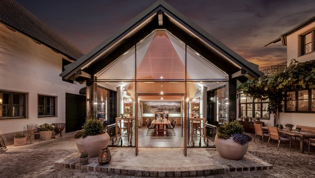 Modern tasting room with glass front and wooden tables, surrounded by traditional buildings at dusk.