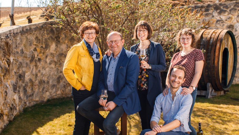 A smiling family poses outdoors in front of a stone wall with a wine barrel in the background.