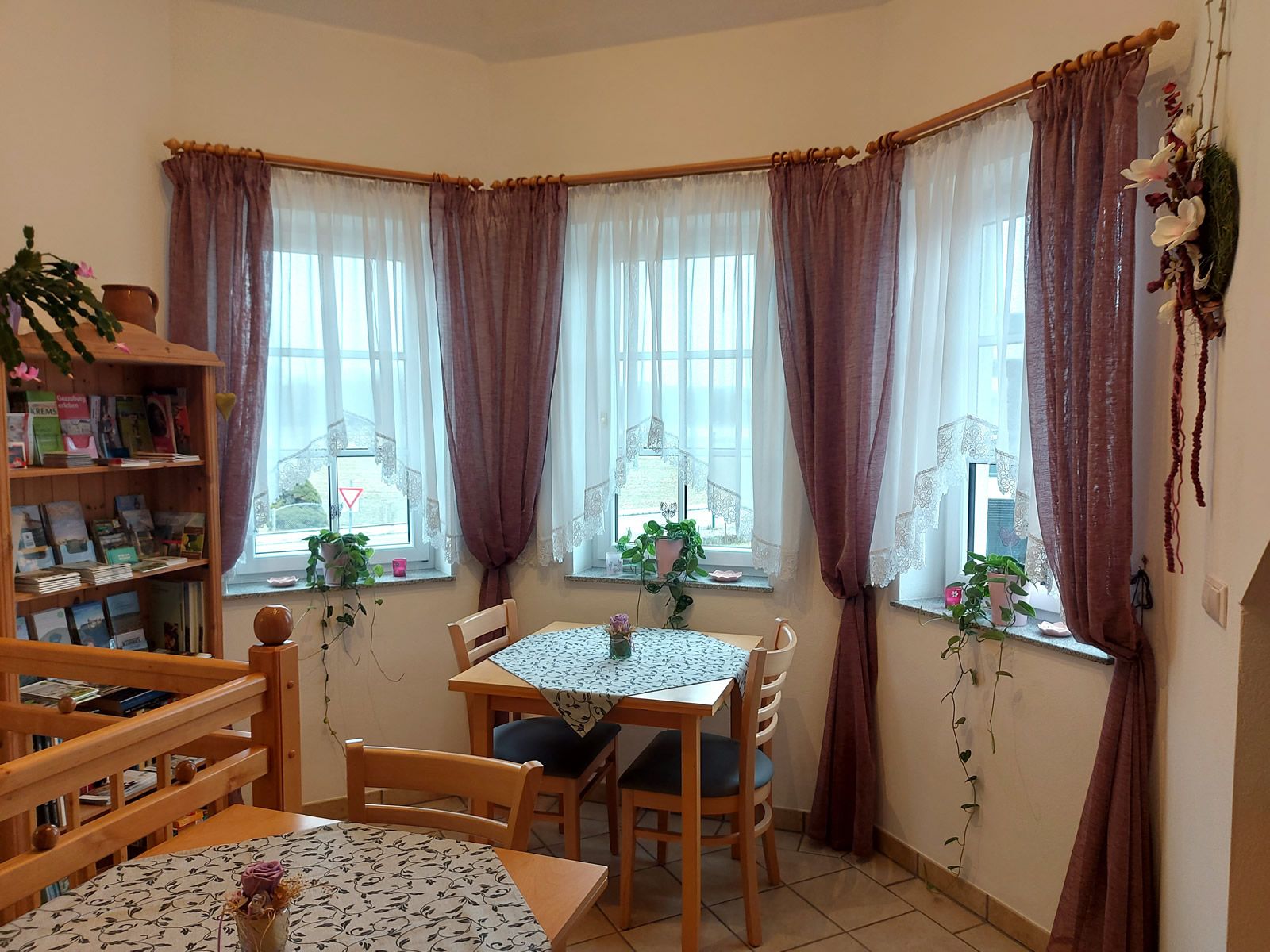 A cozy breakfast room with wooden furniture, tablecloths and plants on the windowsills. Three windows with curtains let in daylight.