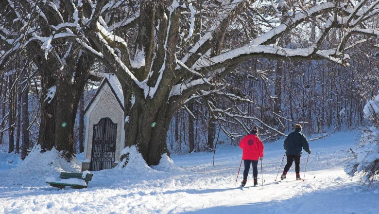 Two cross-country skiers on a snow-covered trail next to a chapel and large trees.