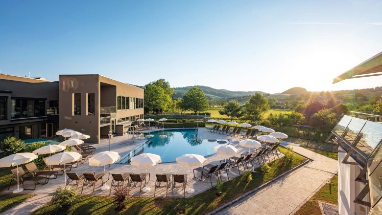 Outdoor area of a hotel with pool, parasols, sun loungers, surrounded by green meadows and trees.