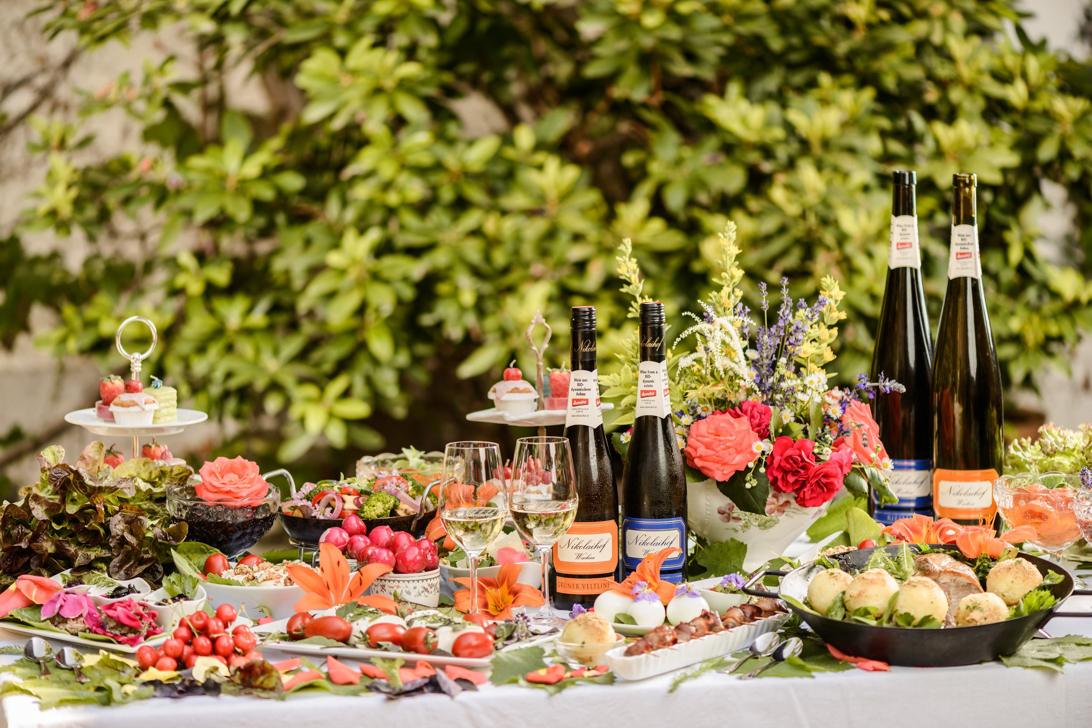 A richly laid outdoor table with bottles of wine, glasses, flowers and various dishes.