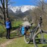A person wearing a helmet stands next to bicycles on a path with snow-covered mountains in the background.
