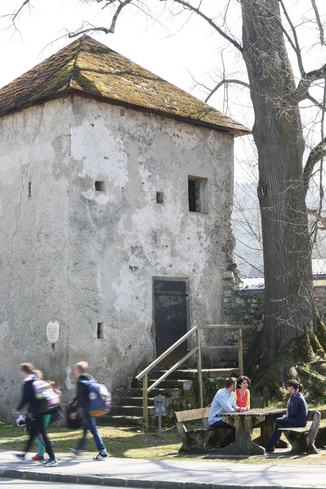 An old tower with a moss-covered roof and a tree next to it. Three people are sitting at a table while two people walk past.