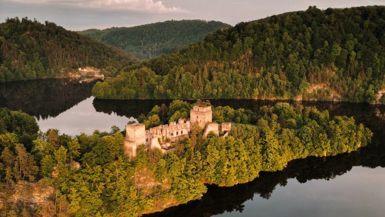 Aerial view of a ruined castle on a wooded peninsula surrounded by a calm lake.