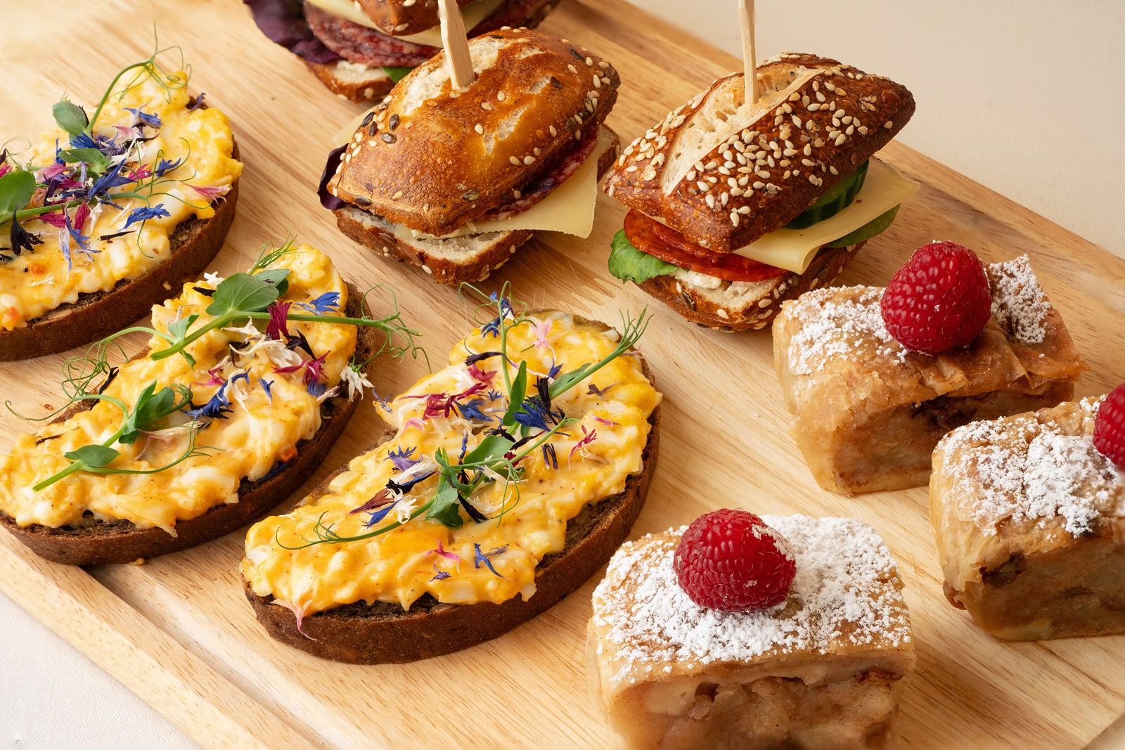 Various snacks on a wooden board, including sandwiches, sandwiches and pastries with raspberries.