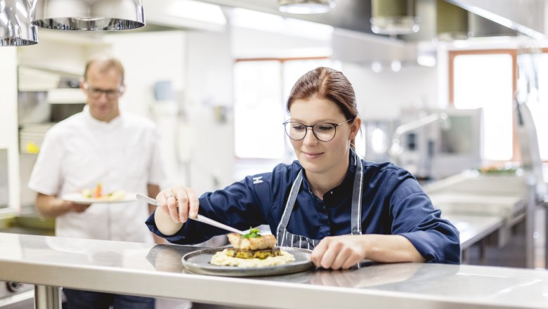 A chef in a restaurant kitchen prepares a dish while a colleague holds another dish in the background.