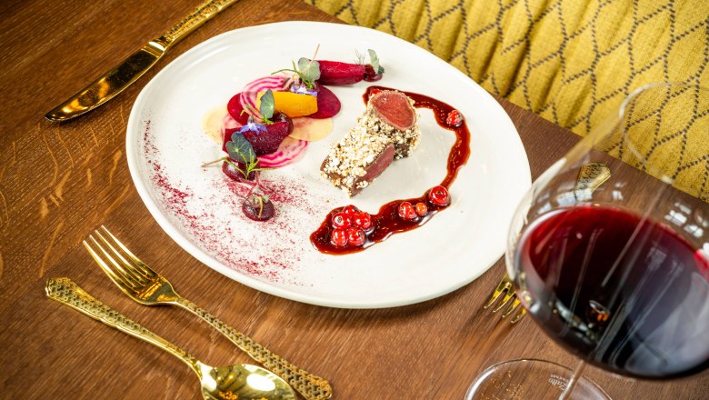 A plate with artfully arranged saddle of venison, side dishes and a red wine sauce, next to a glass of red wine on a wooden table.