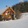 A snow-covered wooden house on the edge of the forest with a blue sky.