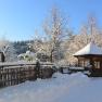 Snow-covered landscape with wooden hut, wooden fence and trees under a blue sky.