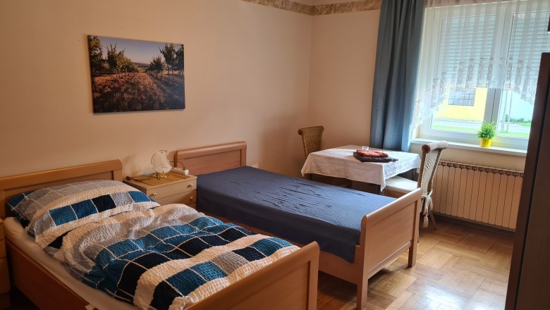 House Waschbergblick, © Wolfgang Greil A simply furnished bedroom with two single beds, a table and chair and a window with curtains.