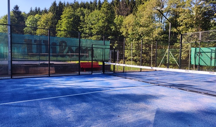 Padel court with blue floor and net, surrounded by trees.