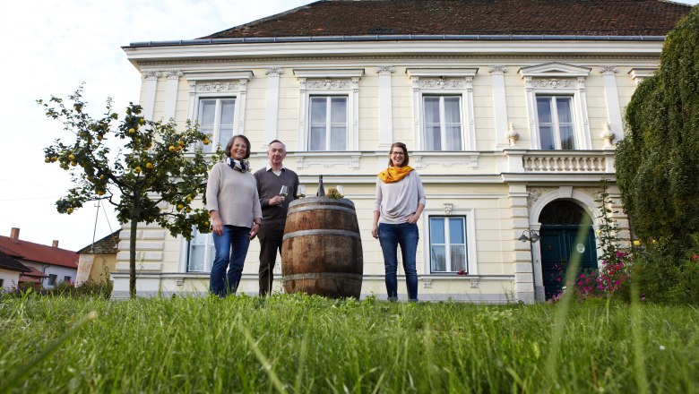 Three people are standing in front of a large yellow building with a barrel in the foreground.